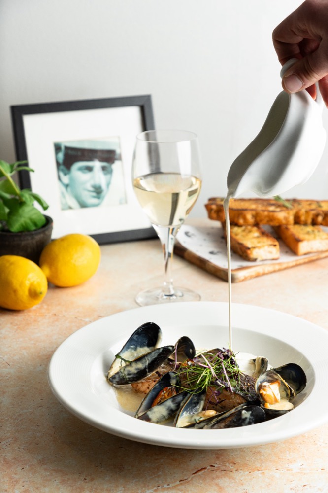 Cream being poured over a bowl of mussels, with a glass of white wine