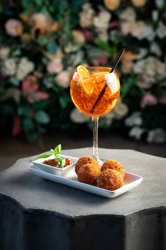 Aperol spritz beside a plate of arancini with dipping sauce, against a floral wall