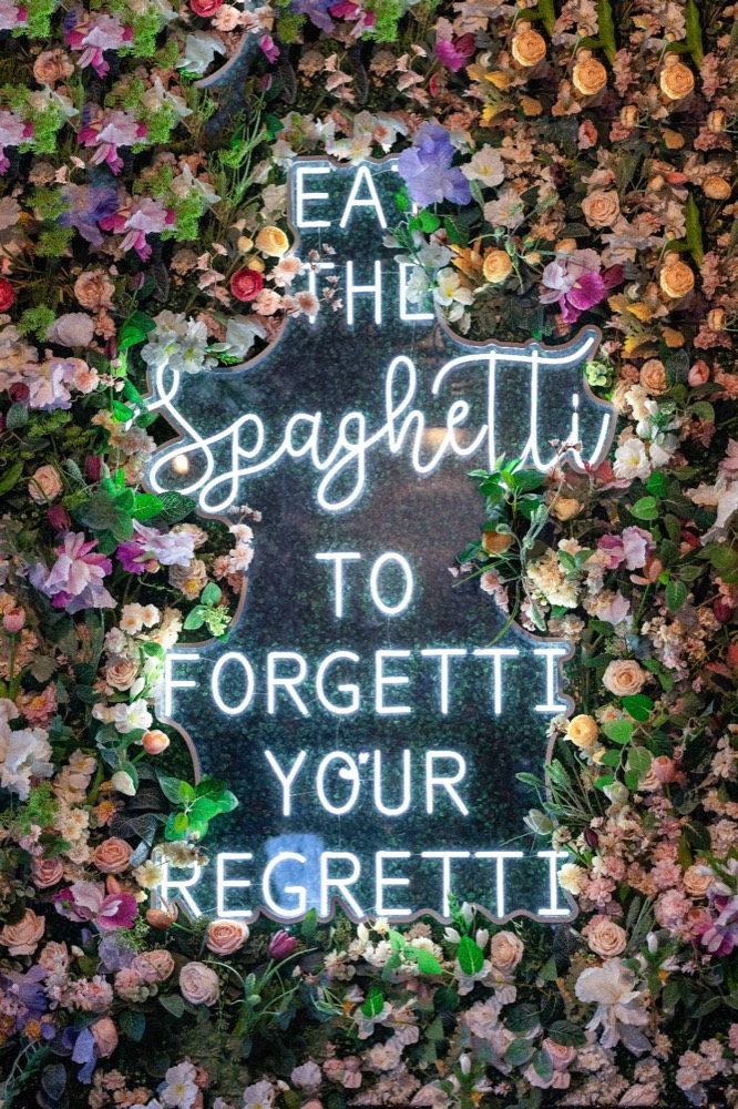 Neon sign reading 'Eat the spaghetti to forgetti your regretti' on a floral wall